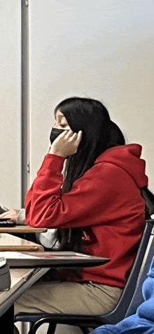 a girl in a red hoodie sits at a desk with a mask on her face