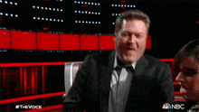 a man in a suit is laughing while sitting next to a woman in a chair with the words #thevoice on the bottom