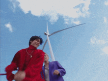 a man in a red jacket is standing next to a man in a purple jacket in front of a windmill