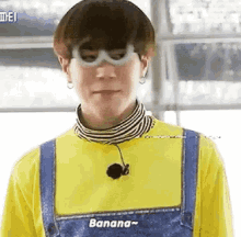 a young man wearing a yellow shirt and blue overalls is wearing a banana mask .