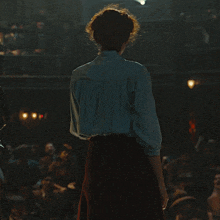 a woman in a blue shirt and red skirt stands in front of a crowd giving a speech