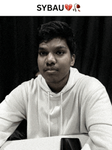 a young man in a white hoodie sits at a table with a broken heart emoji on his face