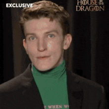 a man is wearing a green turtleneck and a black suit and has a house of the dragon advertisement behind him