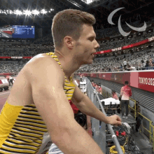 a man in a yellow and black striped tank top stands in front of a sign that says tokyo 2020
