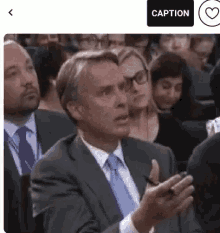 a man in a suit and tie is sitting in a crowd of people and talking to someone .