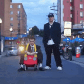 a man in a suit is pushing another man in a red toy car down a street