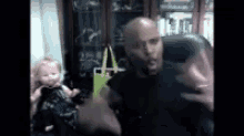 a bald man is sitting in a chair with a doll in the background .