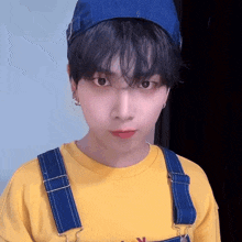 a young man wearing a yellow shirt and blue overalls is looking at the camera