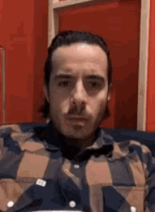 a man in a plaid shirt is sitting on a couch and looking at the camera .