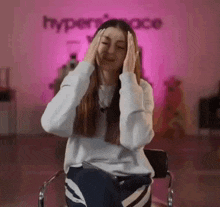 a woman is sitting in a chair with her hands on her head in front of a pink wall .