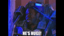 a man is sitting in a chair with a robot on his head and the words `` he 's huge '' .