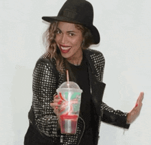 a woman wearing a hat and a studded jacket is holding a plastic cup with a straw .