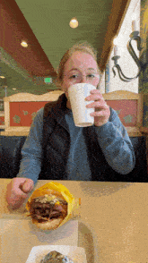 a woman drinking a cup of coffee while eating a hamburger
