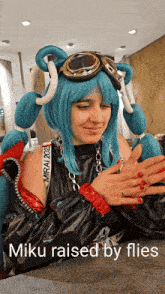 a woman in a blue wig and goggles with the words miku raised by flies below her