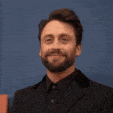 a man with a beard wearing a suit and black shirt smiles for the camera