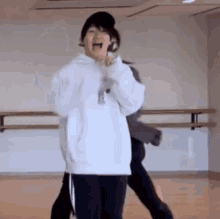 a woman in a white hoodie is dancing in a dance studio with a microphone in her hand .