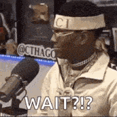 a man wearing a headband and glasses is sitting in front of a microphone and says `` wait ? '' .