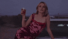 a woman in a pink and red dress is sitting on the hood of a car holding a glass of wine