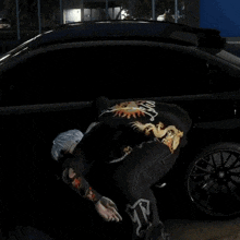 a man in a black shirt with a dragon on the back is squatting next to a black car