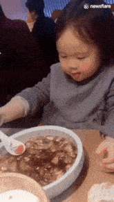 a little girl is sitting at a table with a bowl of food and a spoon in front of her ..