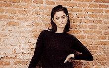 a woman standing in front of a brick wall wearing a black sweater