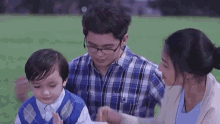 a man and a woman are sitting next to a little boy in a field . the man is wearing glasses .