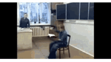 a man is sitting in a chair in a classroom while another man stands in front of a window .