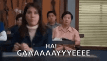 a woman is sitting at a desk in a classroom with other people and making a funny face .