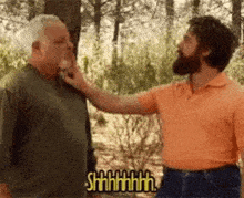 a man with a beard is touching another man 's face in a forest .