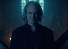 a man with long hair is standing in a dark room with a stained glass window in the background .