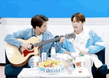 a man playing a guitar next to another man playing a guitar with a table full of bottles of 0