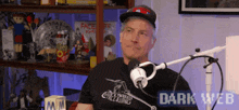 a man is sitting in front of a microphone wearing a hat and a t-shirt that says dark web .