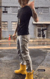 a man wearing a black shirt and yellow rain boots stands in the rain
