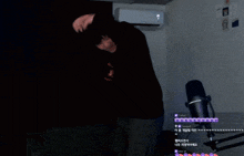 a man in a black hoodie stands in front of a microphone in a dark room with a white air conditioner