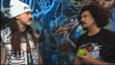 two men are standing in front of a graffiti wall one is wearing a white beanie that says ' alive '