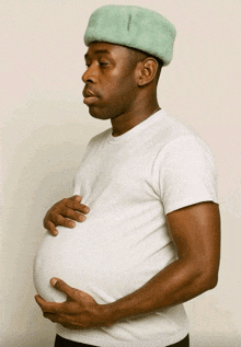 a man wearing a white shirt and a green hat is holding his pregnant belly