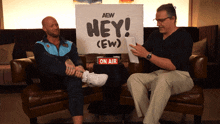 two men are sitting on a couch with a sign that says hey