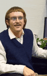 a man wearing glasses and a blue vest looks at the camera