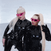two women wearing black latex outfits and pink sunglasses pose for a picture