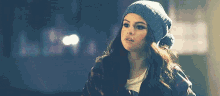 a woman wearing a beanie and a plaid jacket is standing in the dark .