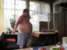 a shirtless man is standing in front of a window in a room with a table and chairs .