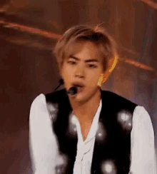 a young man is singing into a microphone while wearing a black vest and white shirt .