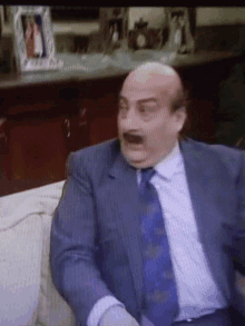 a bald man in a suit and tie is sitting on a couch making a funny face .