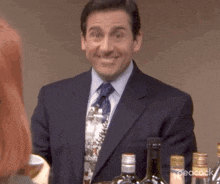 a man in a suit and tie is smiling while standing next to a bottle of wine .