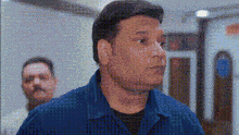 a man in a blue shirt is standing in a hallway with a man in a black shirt behind him .
