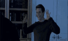 a man in a black sweater is standing in front of a bookshelf with his arms in the air .
