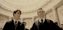 two men in suits and ties stand in a room with columns