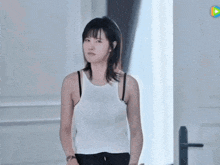 a woman in a white tank top and black bra is standing in front of a window .