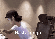 a man wearing a baseball cap is sitting at a desk with the words hasta luego written on the screen .
