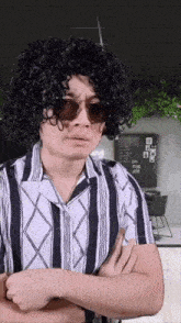 a man with curly hair wearing sunglasses and a striped shirt stands with his arms crossed